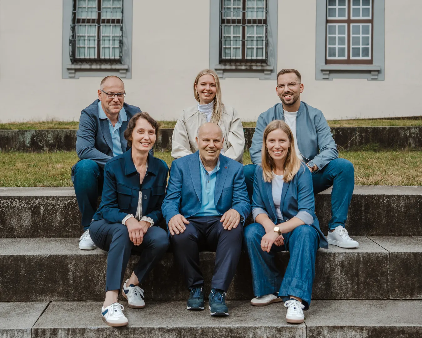Das Fördermittelberater-Team der Deutschen Fördermittelberatung sitzt gemeinsam auf Steinstufen vor einem Gebäude. Die Personen wirken offen, vertrauenswürdig und engagiert.
